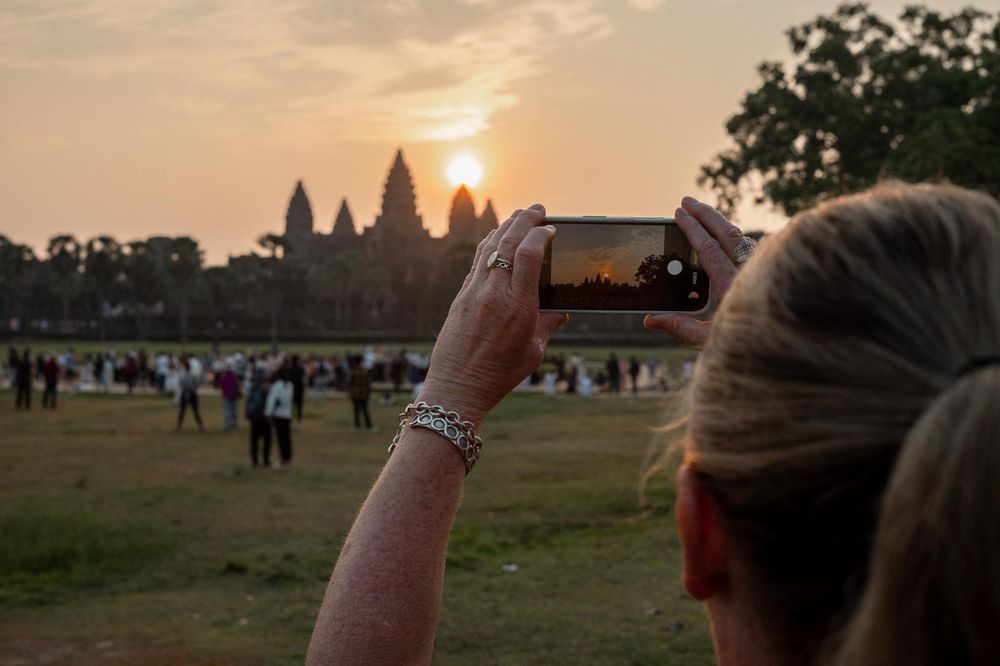 Woman photographing Angkor Wat temple sunrise on smartphone with crowds gathered at the historic Cambodia landmark.