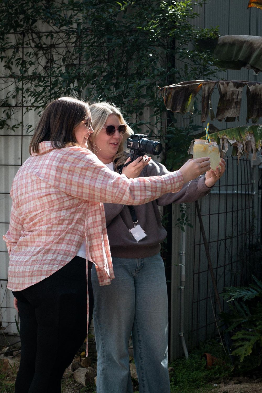 Two women examining plants outdoors, one holding a camera while the other holds a yellow flower pot near a garden fence.