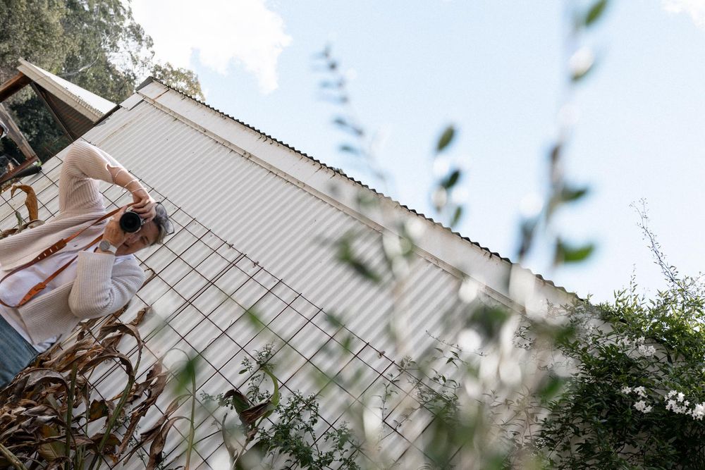 Person in white outfit photographing a white corrugated building surrounded by lush greenery on a sunny day.