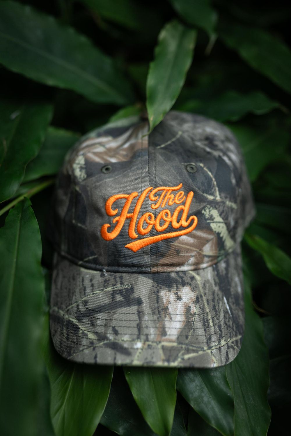 Camouflage baseball cap with orange embroidered The Hood logo resting on tropical green leaves.