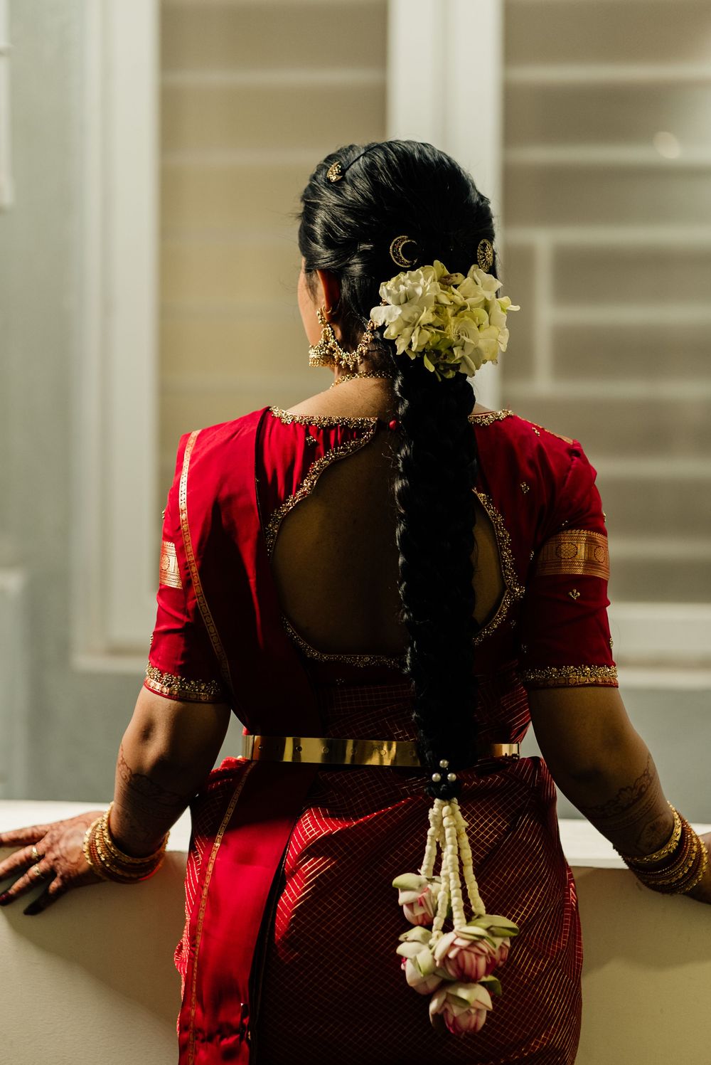 Indian bride in red saree with intricate braid adorned with white flowers and gold jewelry at wedding.