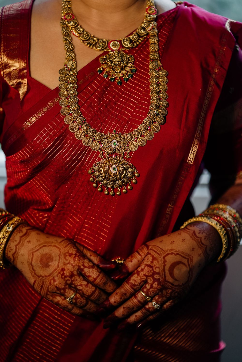 Indian bride in red saree with intricate gold temple jewelry and henna-adorned hands clasped together.