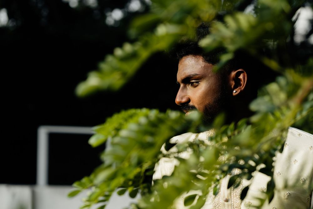 A pensive South Asian man in white attire partially obscured by lush green fern leaves in soft natural light.