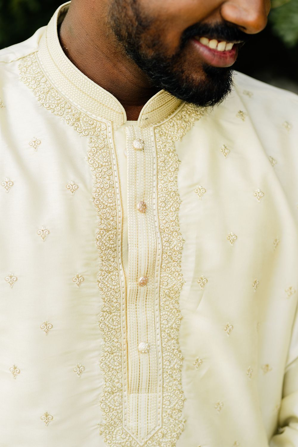 Man smiling in cream embroidered kurta with intricate gold threadwork and decorative buttons at the neckline.