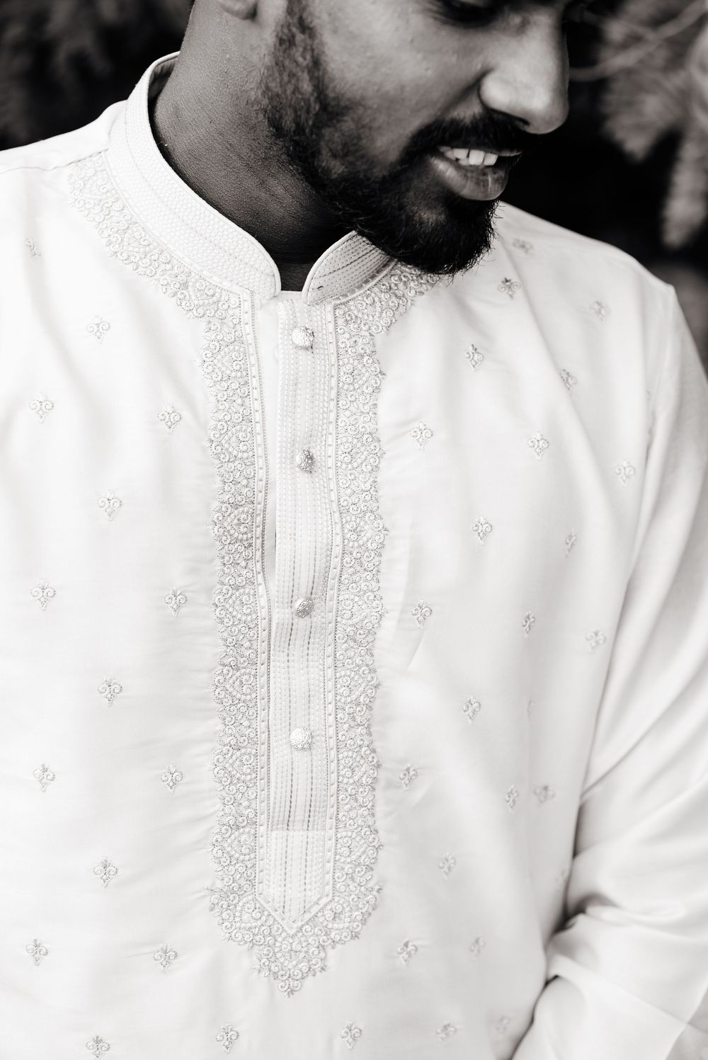 Black and white portrait of a smiling bearded man wearing an embroidered white kurta with intricate detailing.