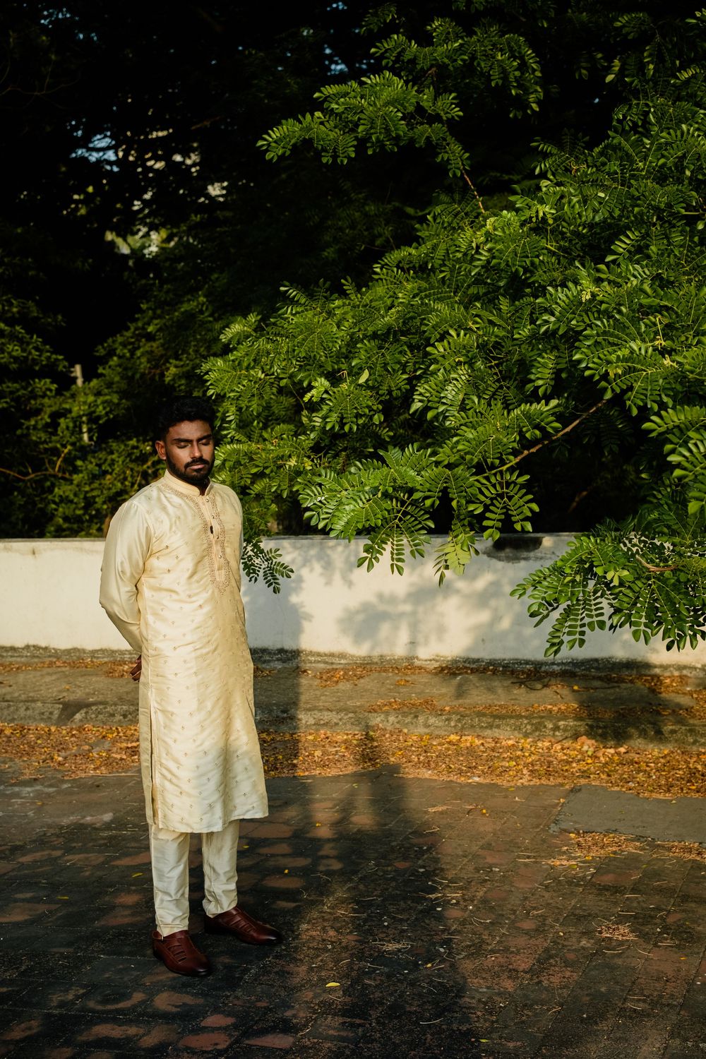 Indian groom in traditional cream kurta pajama standing outdoors near lush green trees.