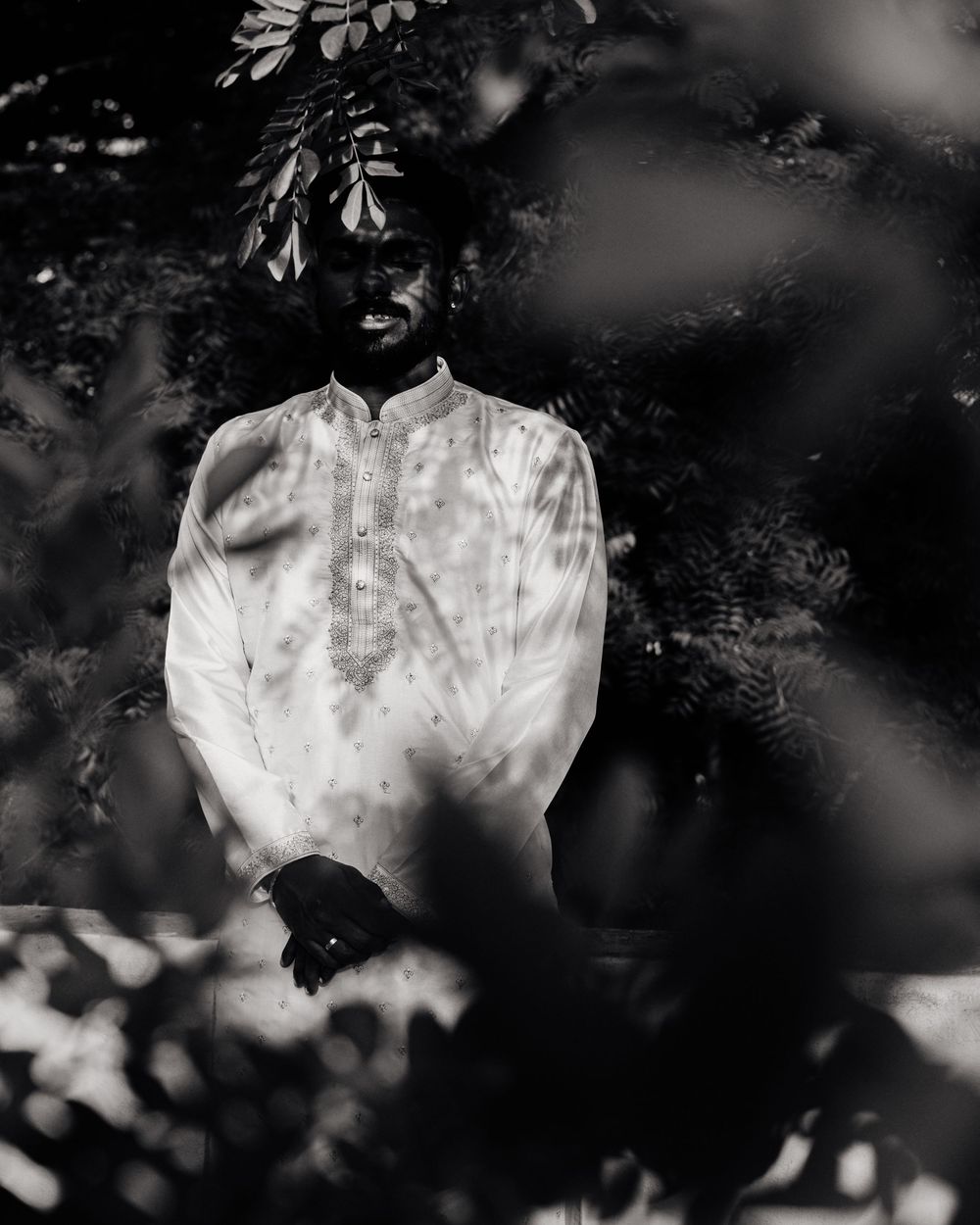 Black and white portrait of Indian groom in white kurta framed by leafy branches.