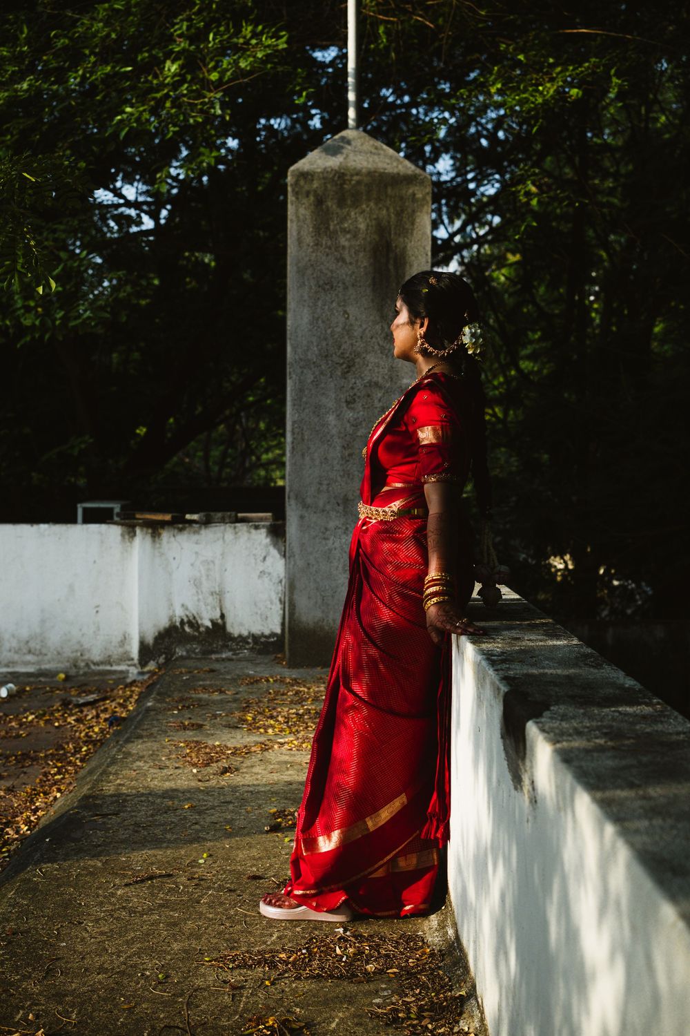 Indian bride in elegant red silk saree with gold border standing outdoors in sunlight.