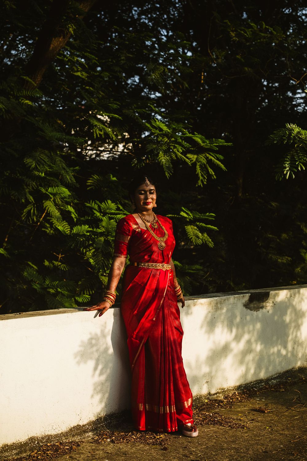 A bride in a red saree with gold jewelry poses against a white wall surrounded by lush green trees.