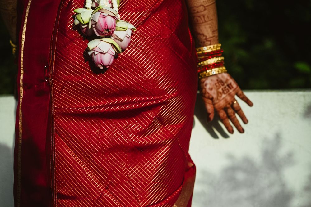 Close-up of a bride in a red saree with gold bangles, henna-adorned hand, and pink lotus flower decorations.