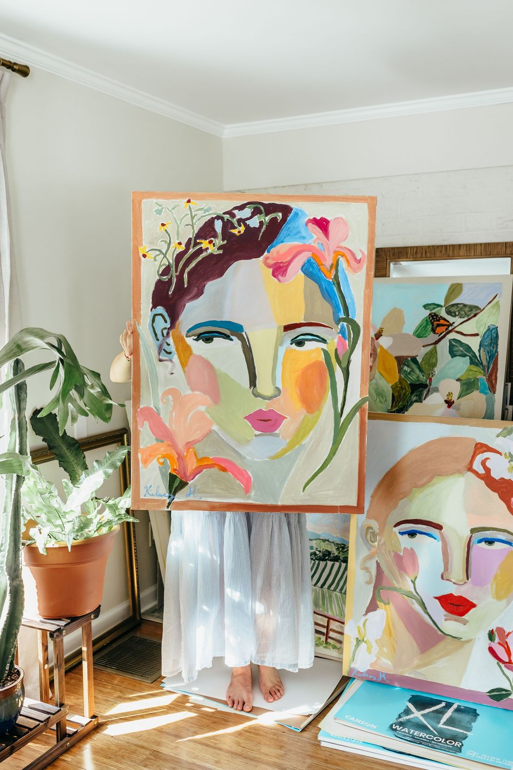 Artist holding a colorful abstract portrait painting in a bright studio surrounded by more face paintings and plants.