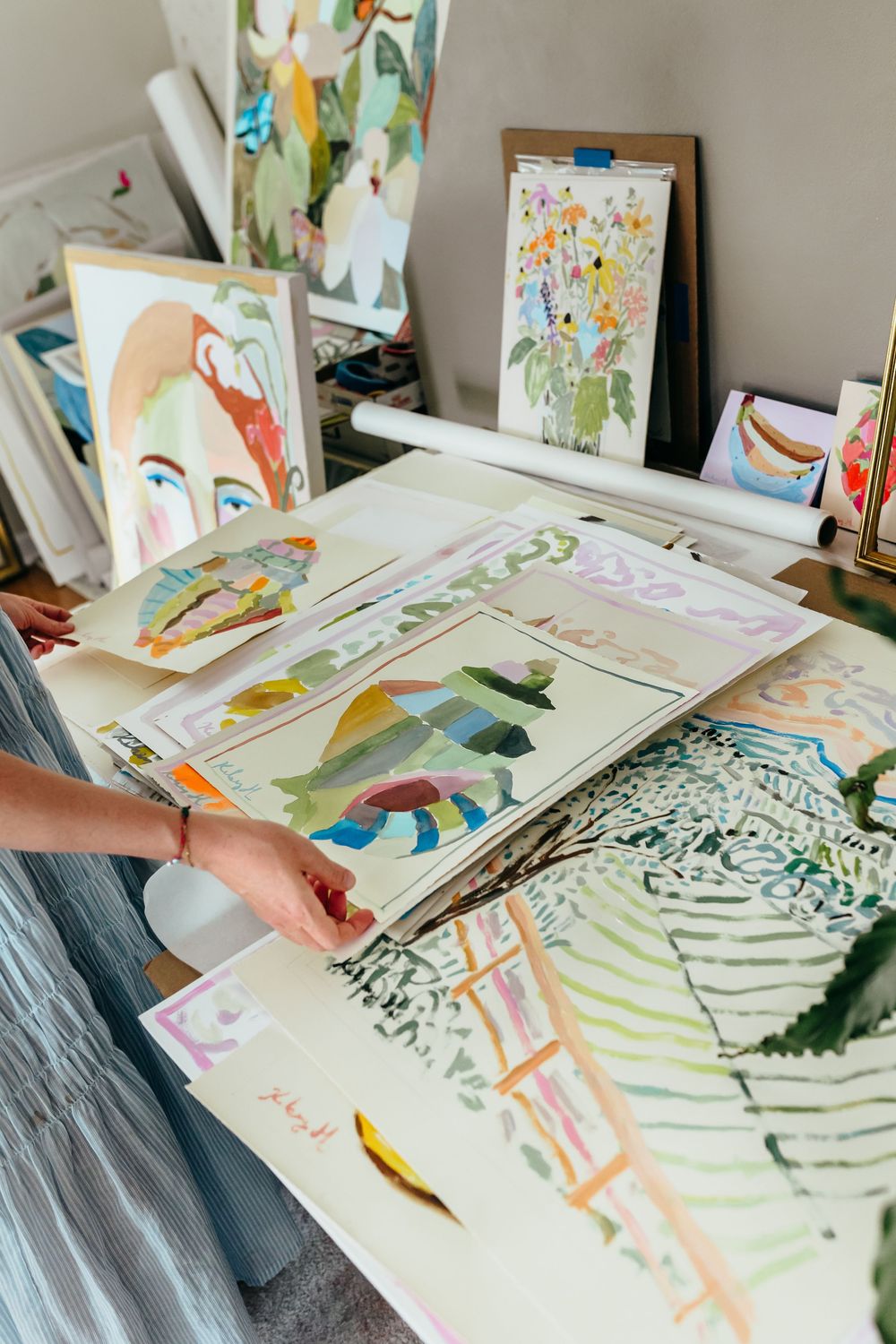 Artist arranging colorful paintings and sketches on a studio worktable surrounded by vibrant artwork on easels.