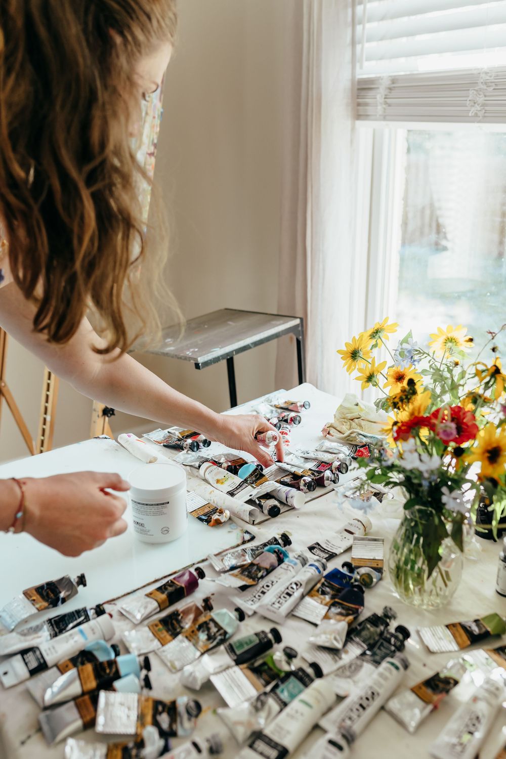 Artist arranging colorful paint tubes on a white table with a vase of sunflowers by a bright window.