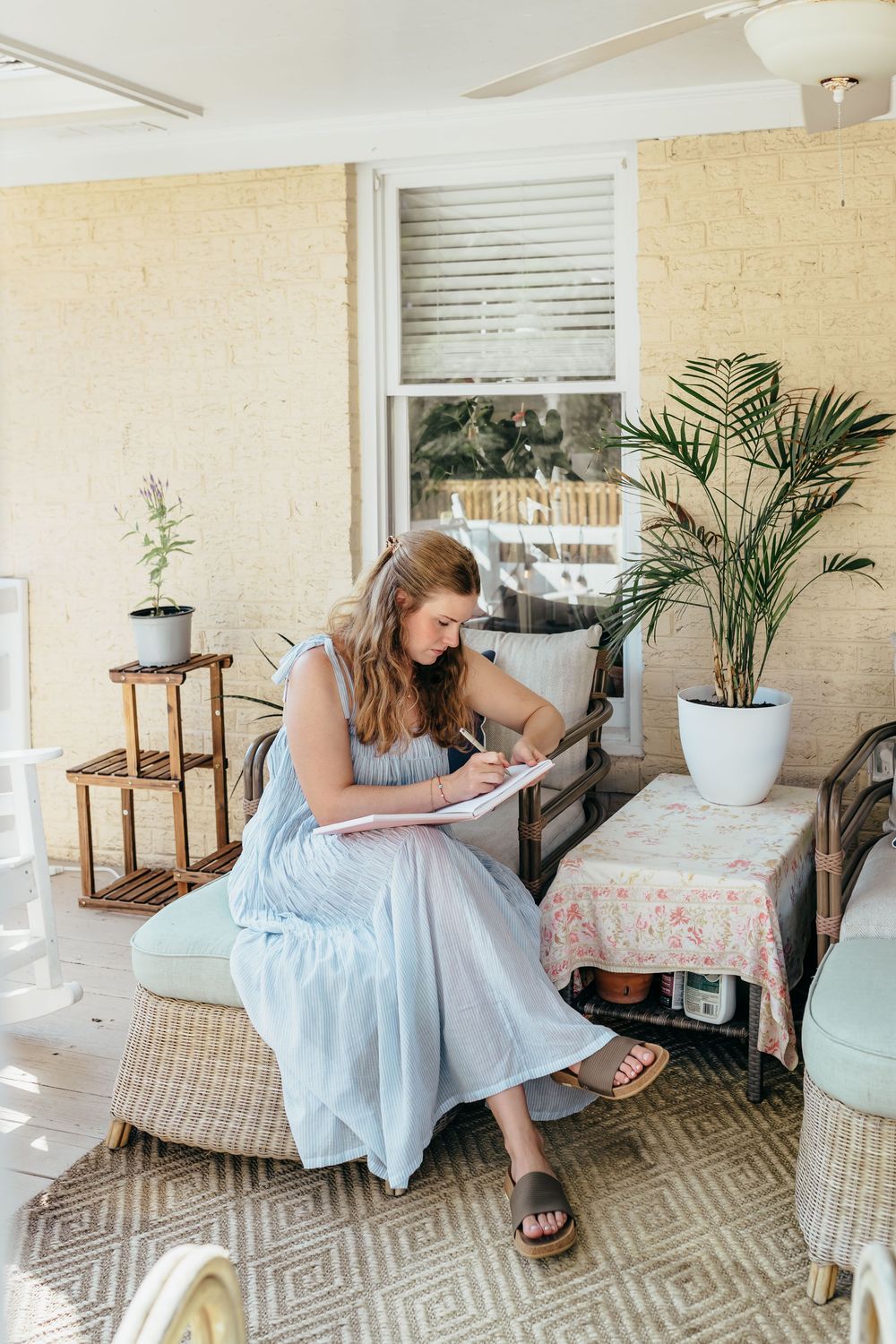 Woman in light blue dress writing in notebook while sitting on wicker chair on a sunlit porch with tropical plants.