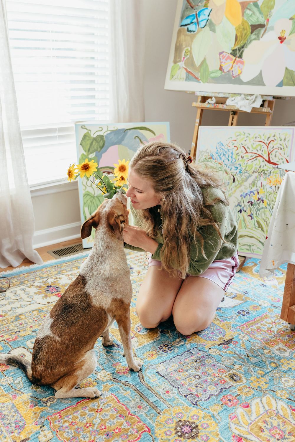 Artist kneeling on colorful rug with small dog in art studio surrounded by vibrant paintings on easels.