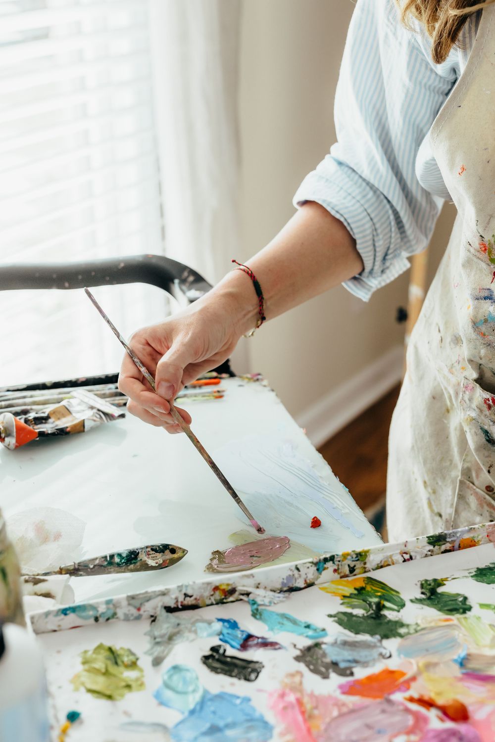 Artist mixing paint colors on a palette with a fine brush in a bright studio, wearing a paint-splattered apron.