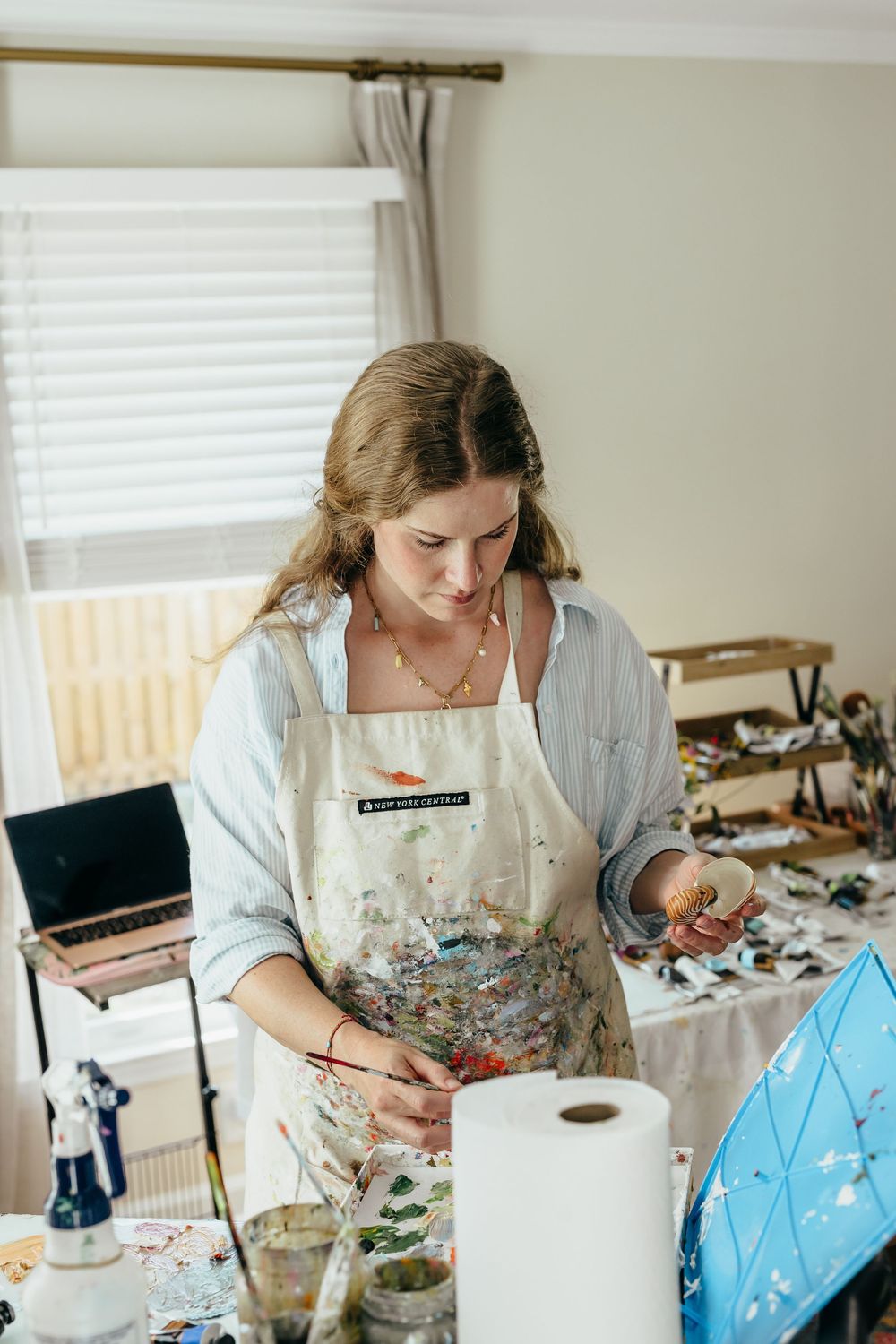 Artist in paint-stained apron works at a cluttered creative studio workspace with art supplies and a laptop.