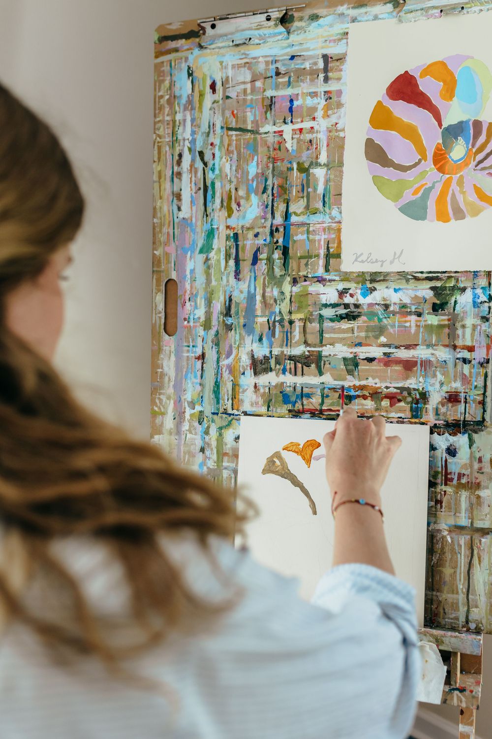 Artist painting a butterfly on canvas at an easel with a colorful abstract palette board in a bright studio.