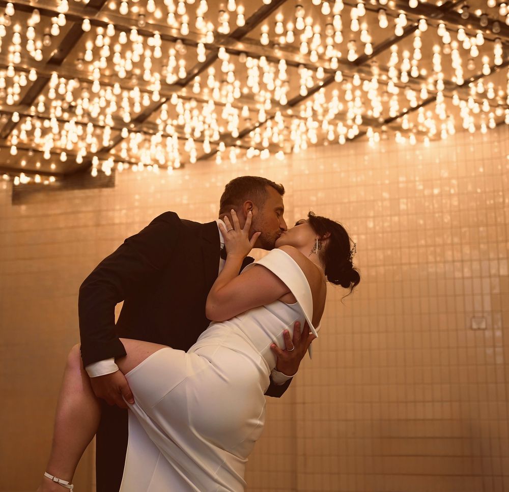 A couple shares a romantic kiss, the groom lifting the bride in a white dress under warm string lights.