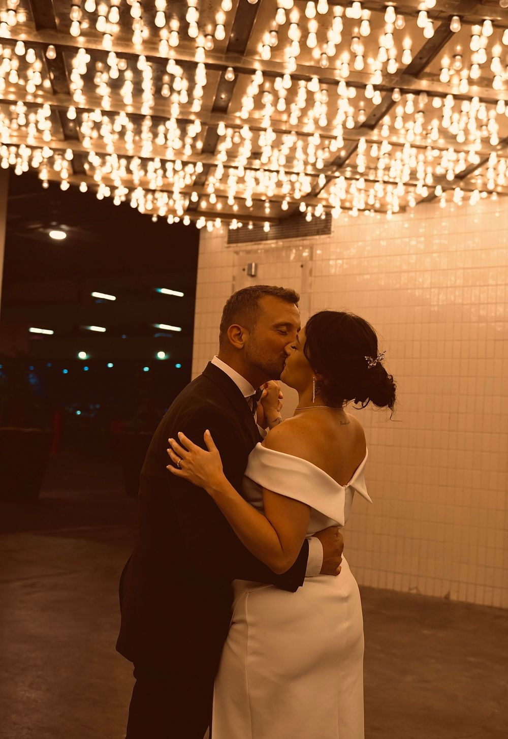 Bride and groom share a romantic first dance kiss under a ceiling of glowing Edison bulb lights at their wedding reception.