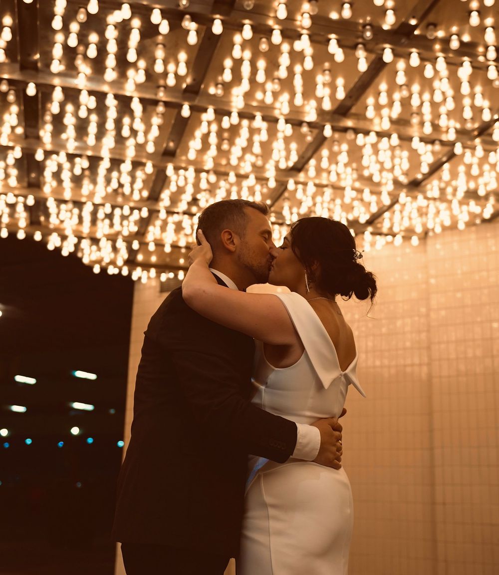 Couple sharing romantic first dance kiss under warm string lights at elegant wedding reception.