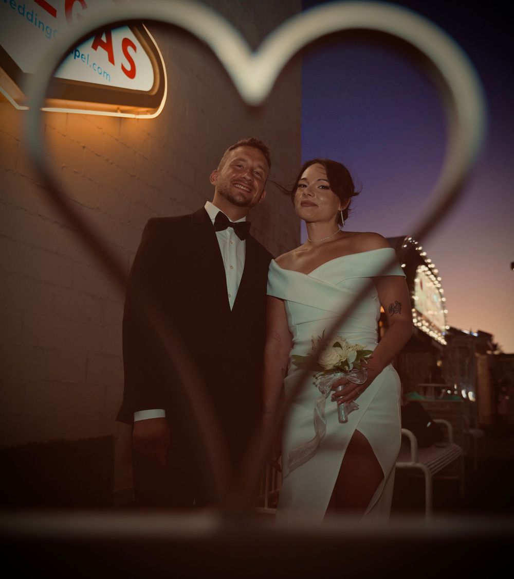Newlywed couple in wedding attire framed by a heart-shaped bokeh at dusk near a Las Vegas neon sign.