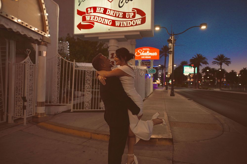 Couple sharing a romantic kiss outside a 24-hour drive-up wedding window chapel in Las Vegas at dusk.