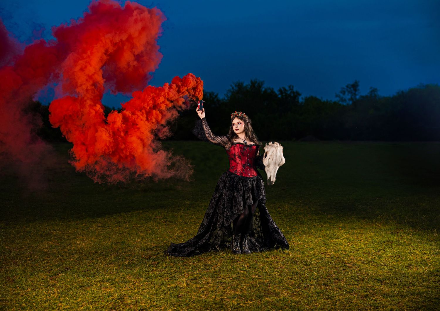 Woman in black and red gown holds mirror amid dramatic red smoke clouds on green field.