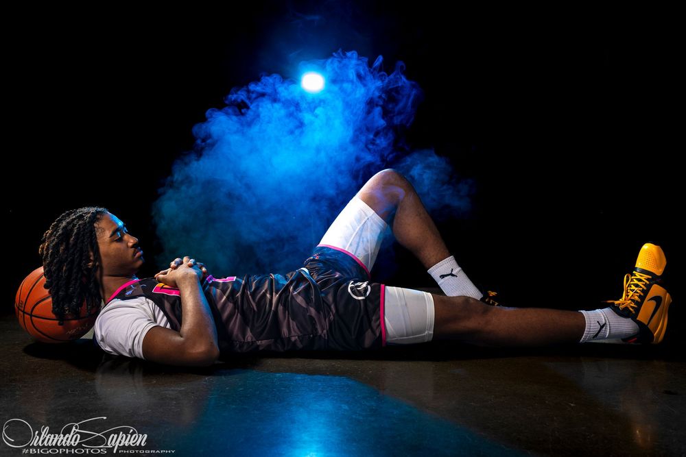 Basketball player in camo shorts poses on floor with ball under blue smoke and dramatic studio lighting.
