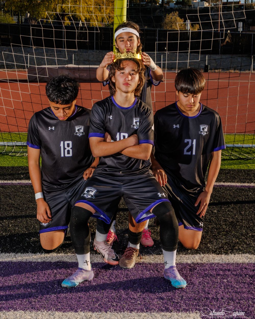 Four youth soccer players in black uniforms pose dramatically on a field in front of a goal net at night.