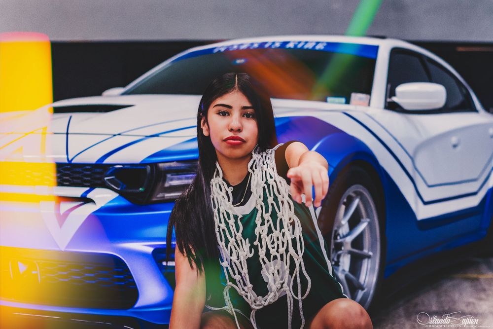 Young woman with beaded necklaces poses in front of a blue Ford Mustang mural outdoors.