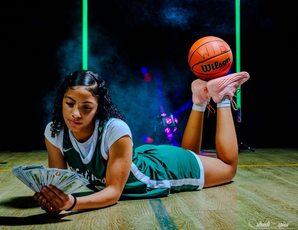 Female basketball player poses on court floor holding cash fan with Wilson basketball balanced on foot.