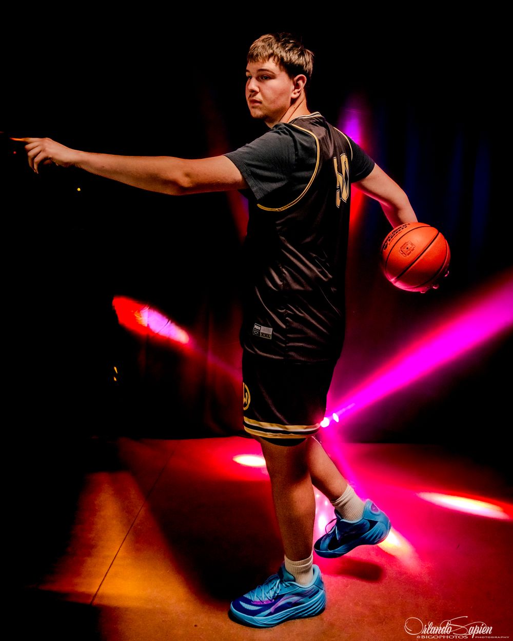 Young basketball player in black jersey dribbles under dramatic pink and red stage lights wearing blue sneakers.