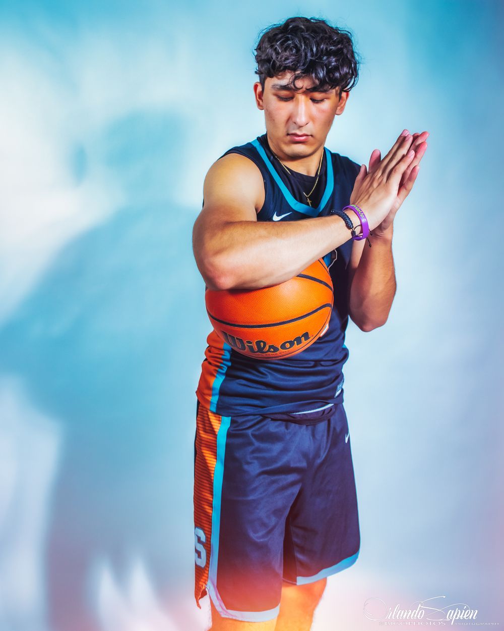 Young male basketball player in navy Nike uniform holds Wilson basketball in dramatic blue-tinted studio lighting.