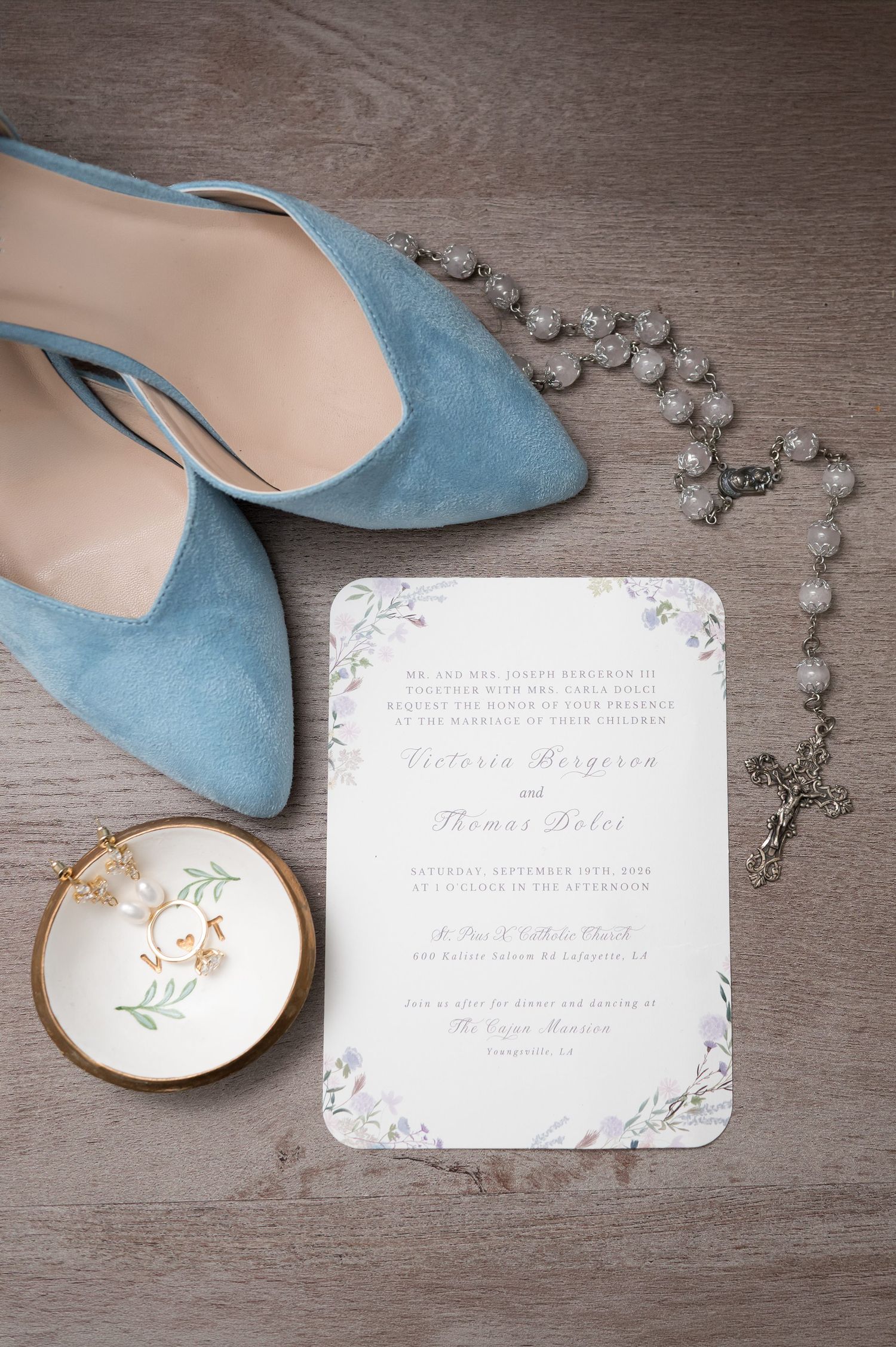 Blue suede wedding heels beside a floral invitation, rosary beads, and a ring dish with pearl earrings on gray wood.