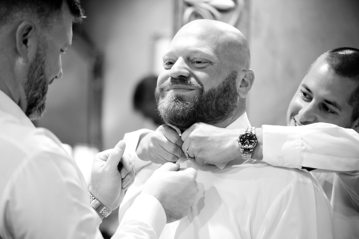 Black and white photo of a bald bearded groom getting his tie adjusted by two groomsmen before wedding.