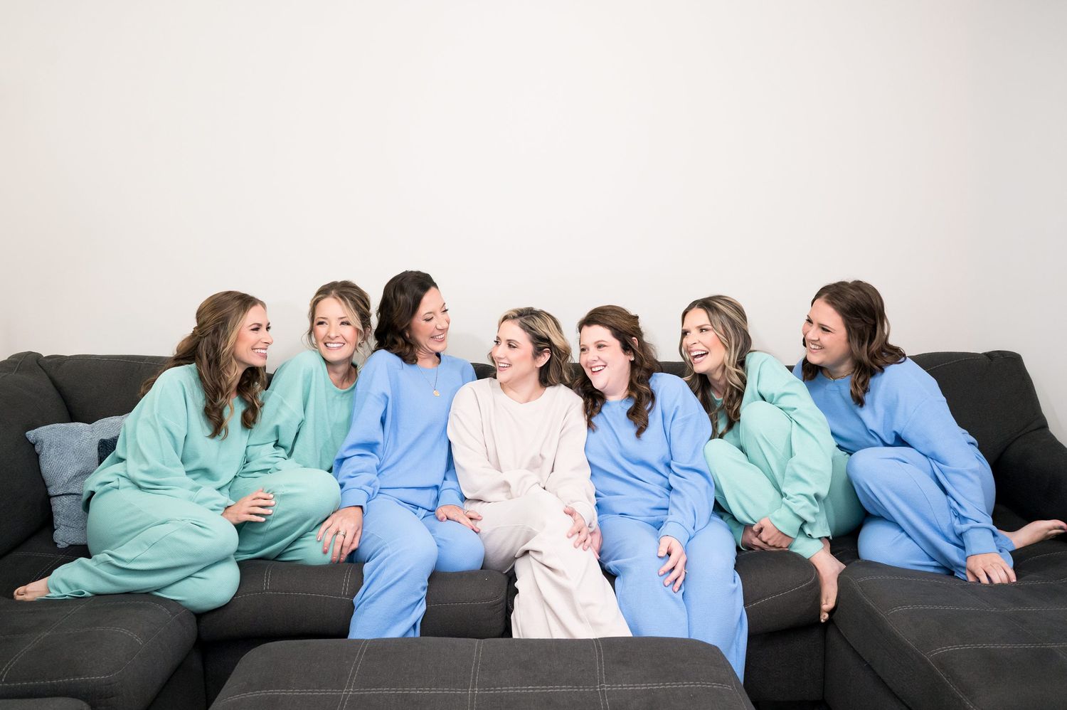 Seven women in matching pastel pajamas smiling and laughing together on a gray couch at a bridal party gathering.