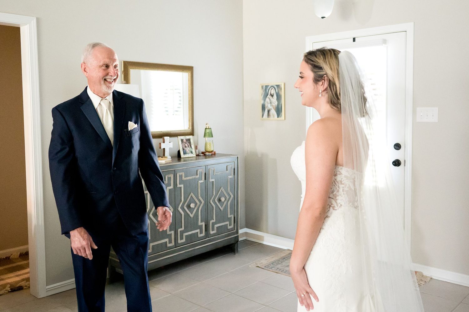 Father smiles at bride in white wedding gown and veil during emotional first look moment inside a home.