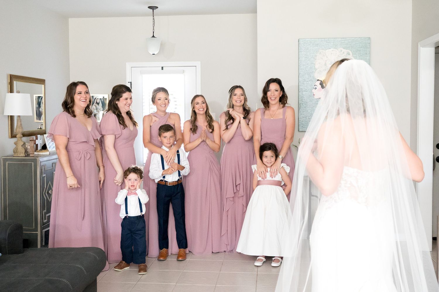 Bridesmaids in dusty rose dresses react with joy as bride reveals her wedding gown.