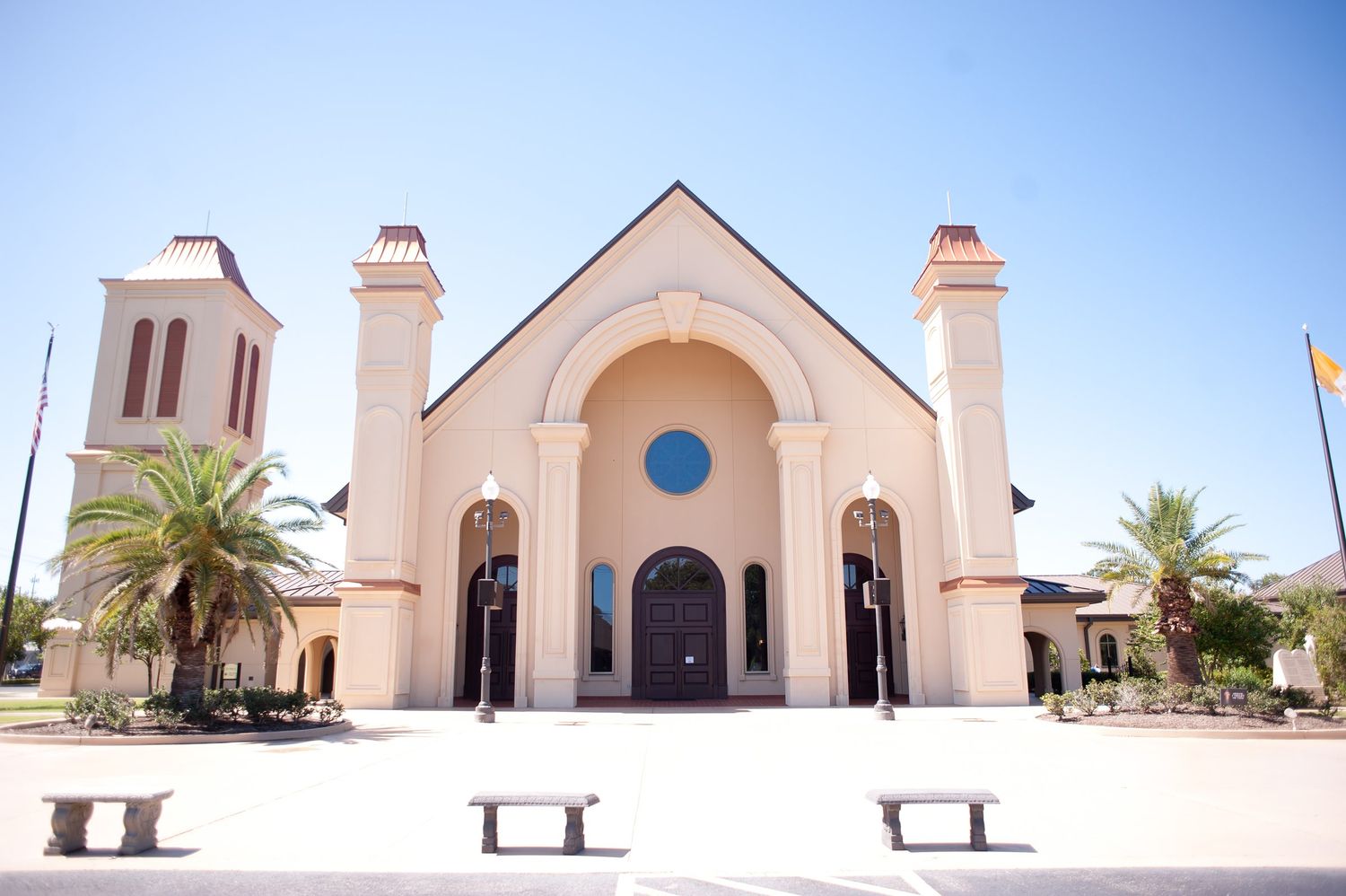 Elegant beige church with twin towers, arched entrance, blue rose window, and palm trees under a clear blue sky.