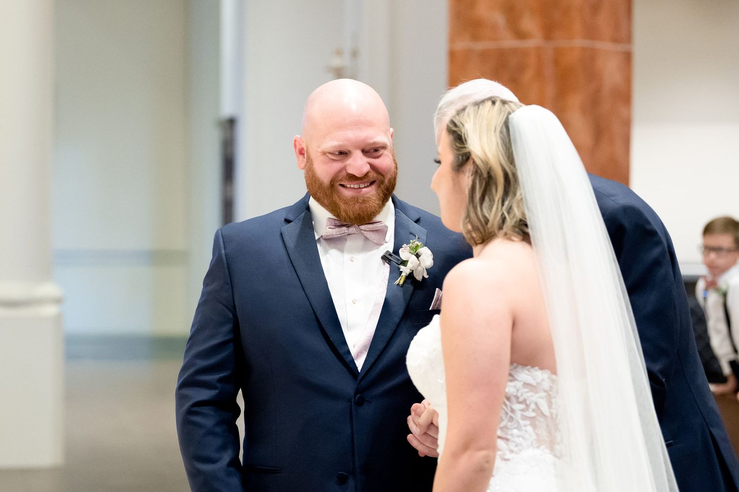 Groom in navy suit smiling joyfully at bride in white lace gown and veil during wedding ceremony.