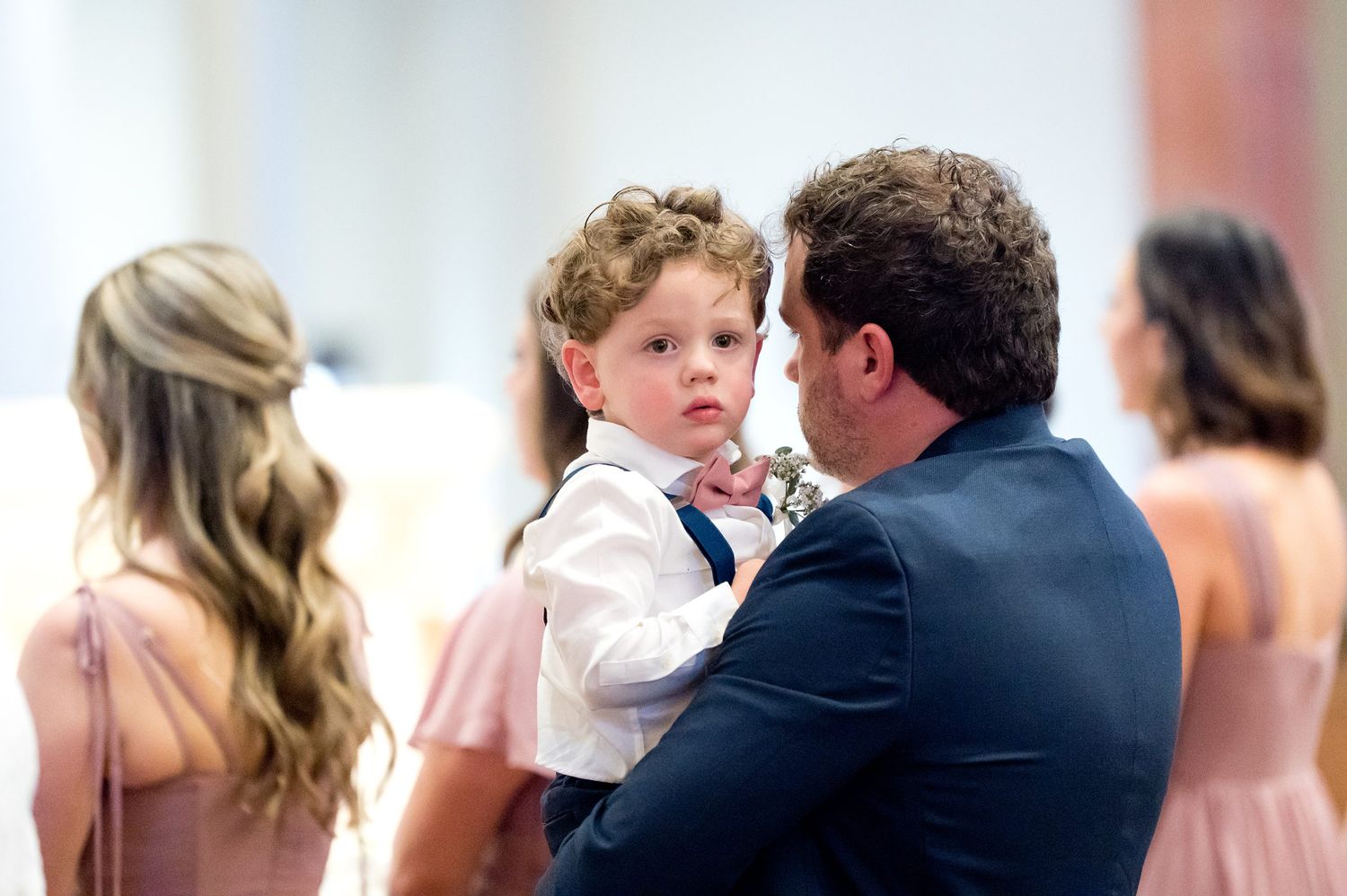A man in a navy suit holds a young boy in a white shirt at a wedding ceremony with bridesmaids in pink.