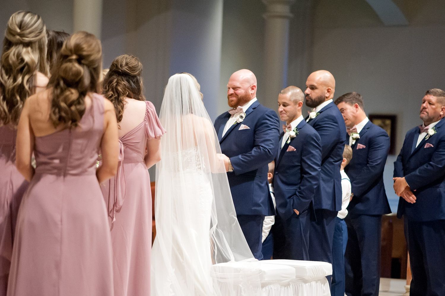 Wedding ceremony with bridesmaids in pink gowns and groomsmen in navy suits facing the bride at the altar.