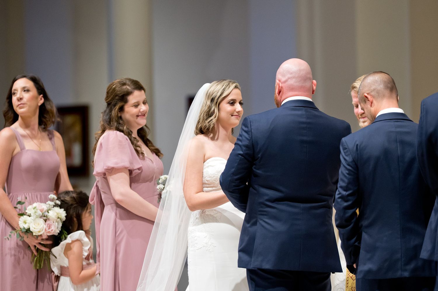 Bride in white gown and veil smiles during wedding ceremony with bridesmaids in pink dresses nearby.