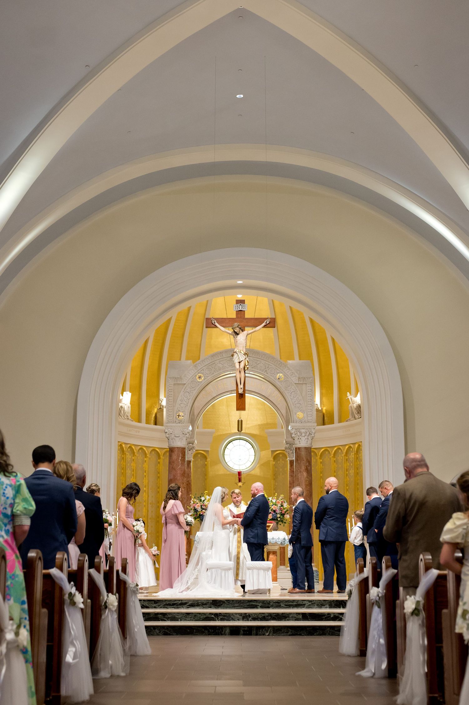 A wedding ceremony at a Catholic church with a crucifix altar, bride and groom at the altar, guests seated in pews.