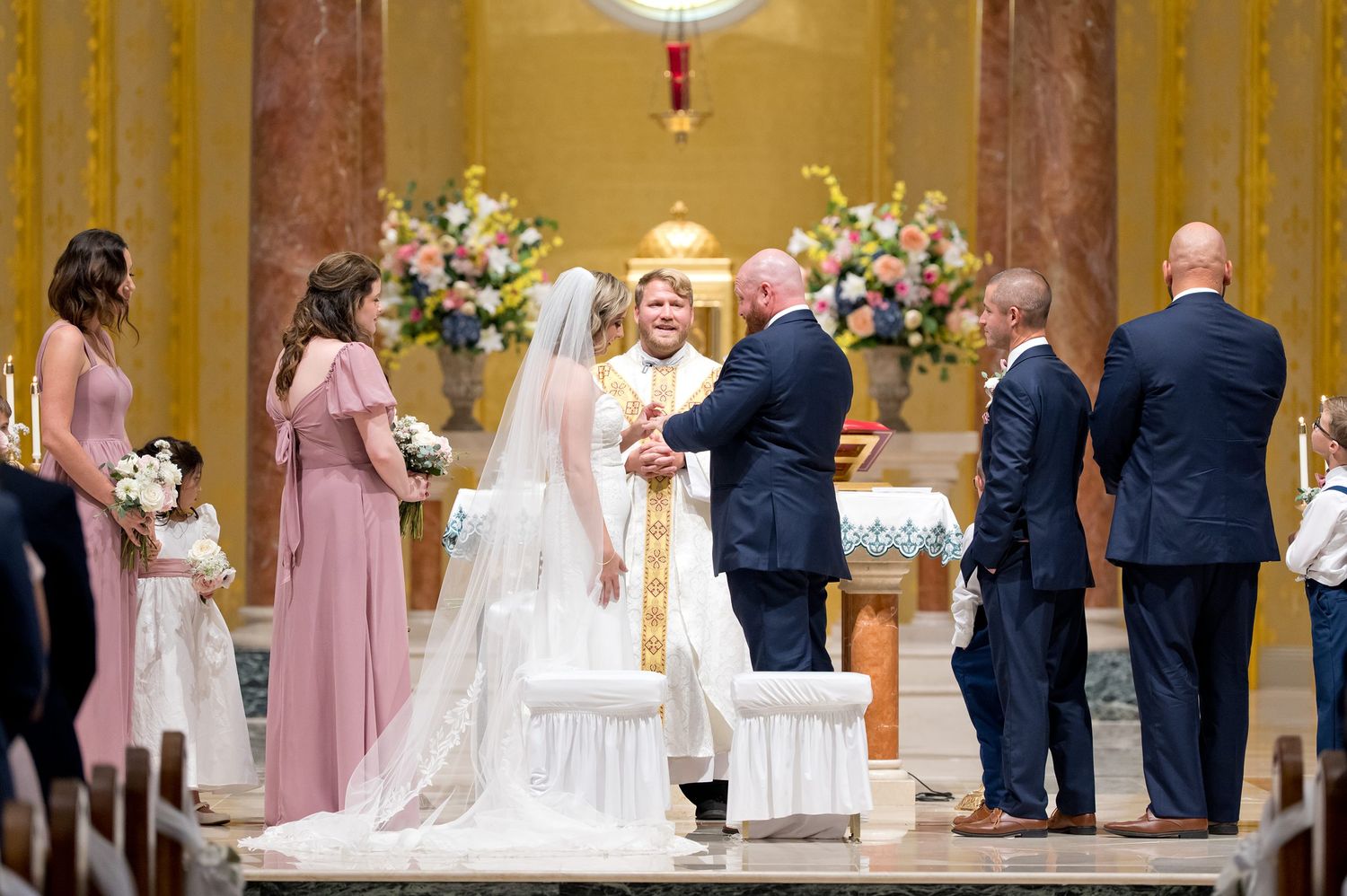 Bride and groom exchange vows at ornate church altar surrounded by wedding party during Catholic ceremony.