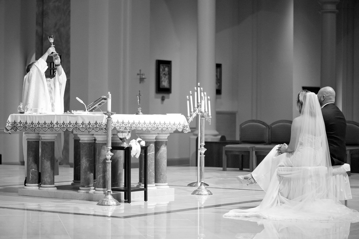Black and white photo of a bride and groom kneeling before a priest at a Catholic church wedding ceremony altar.