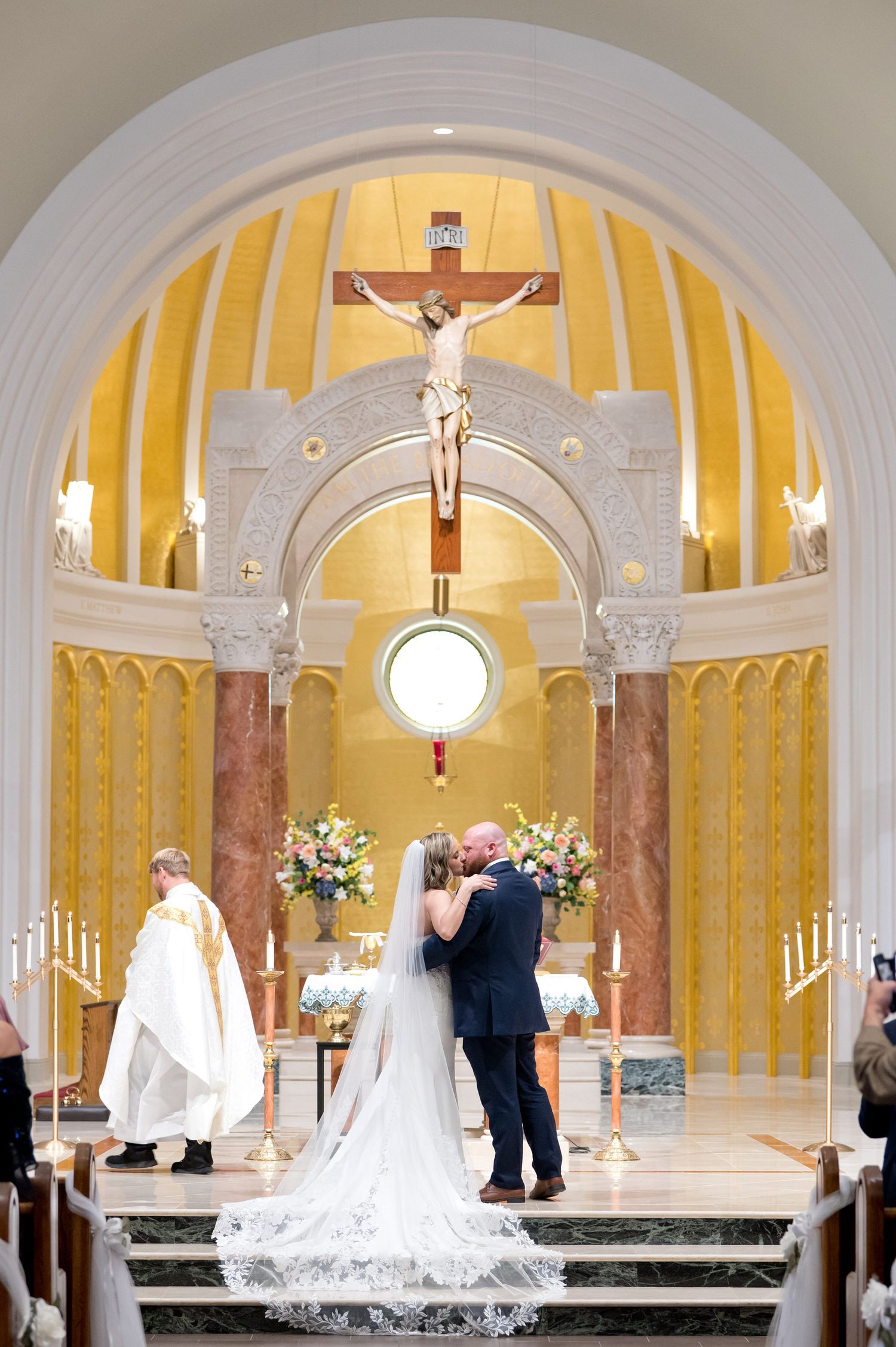 Bride and groom share first kiss at ornate Catholic church altar beneath a crucifix during wedding ceremony.
