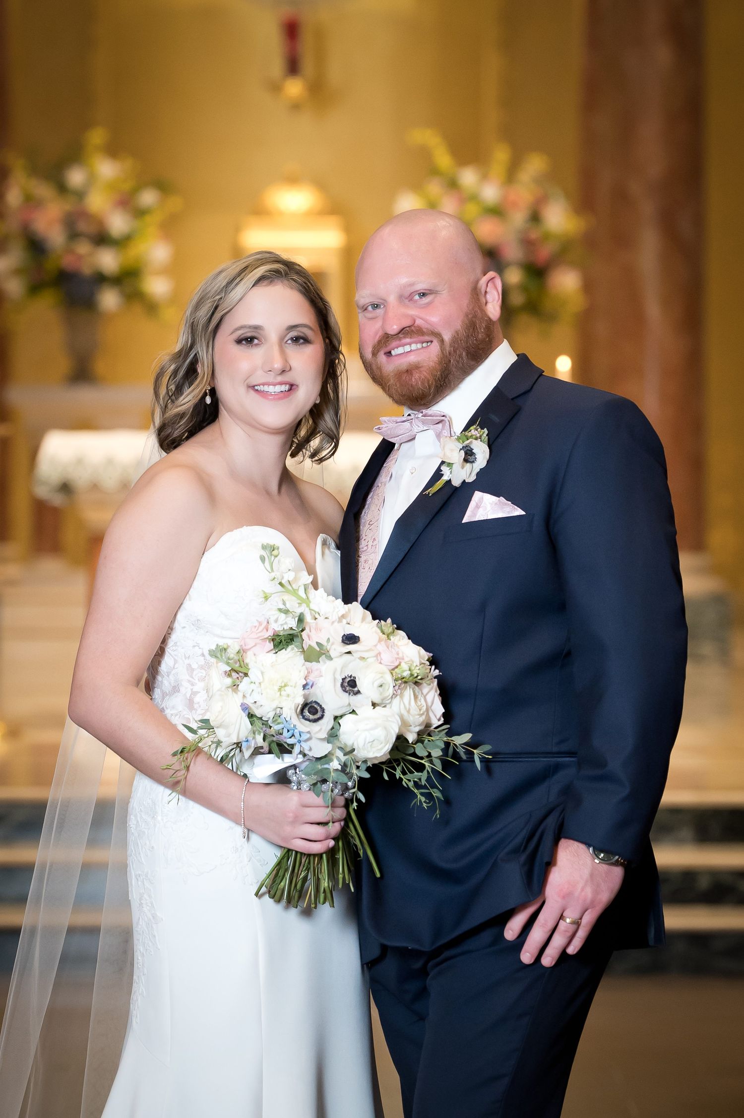 Smiling bride and groom pose together in a church, she holds a white floral bouquet, he wears a navy suit with pink bowtie.