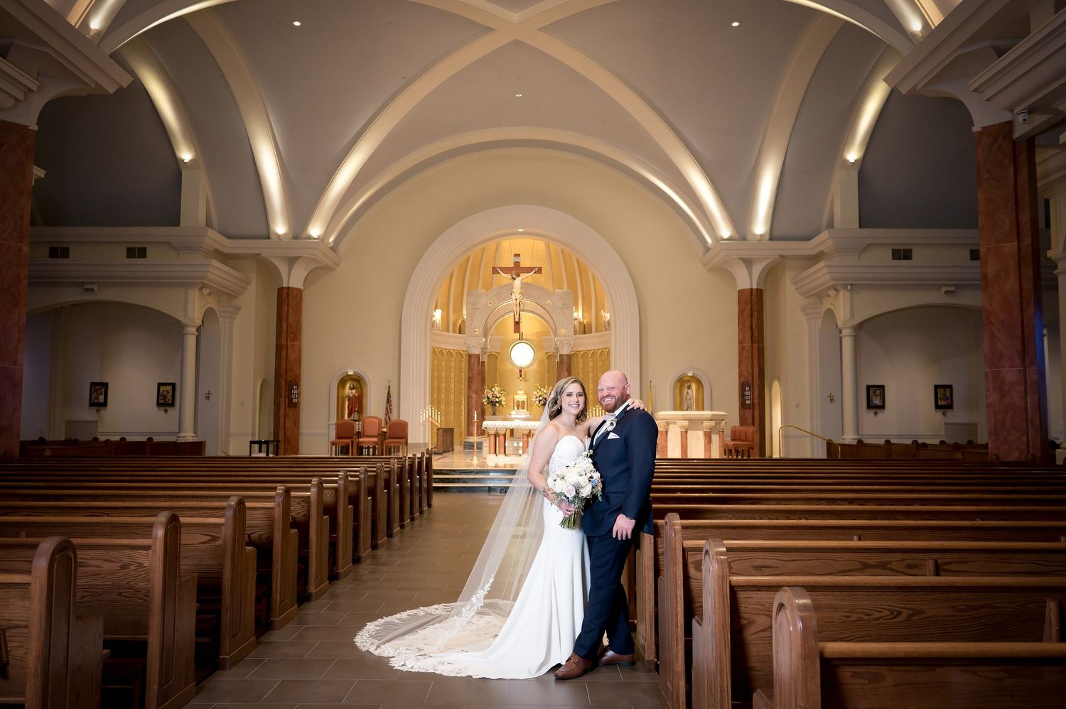 Newlyweds pose in elegant church aisle with vaulted ceilings and wooden pews after wedding ceremony.